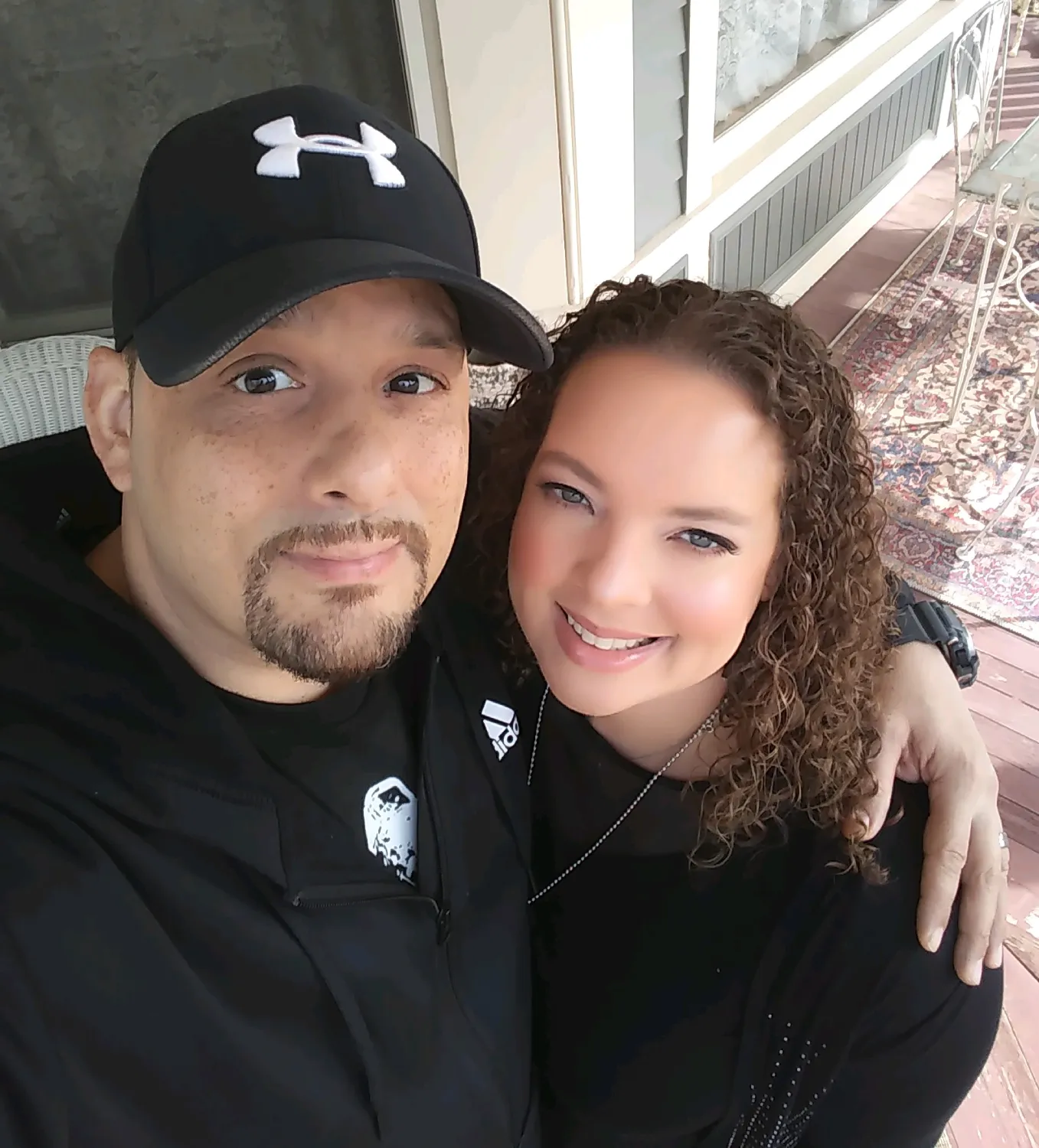 Portrait of a smiling couple sitting together on a porch. One person wears a black baseball cap and black jacket, while the other has curly hair and wears a black top. They are embracing and looking at the camera, with outdoor seating and a patterned rug visible in the background.